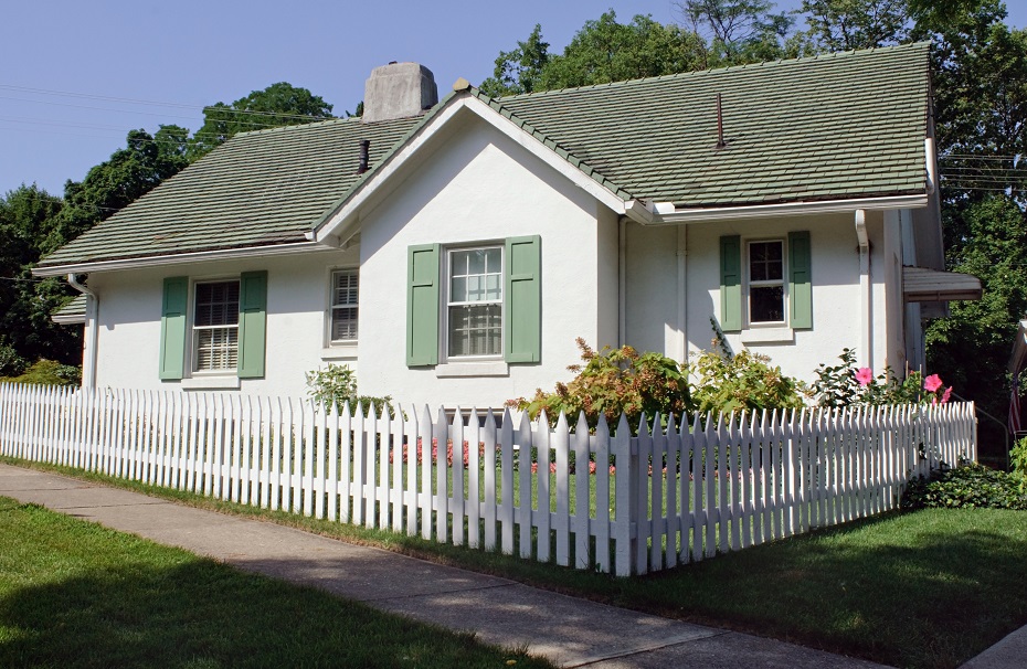 Small house with a sloped roof and white picket fence. It looks wholesome and pleasant. 